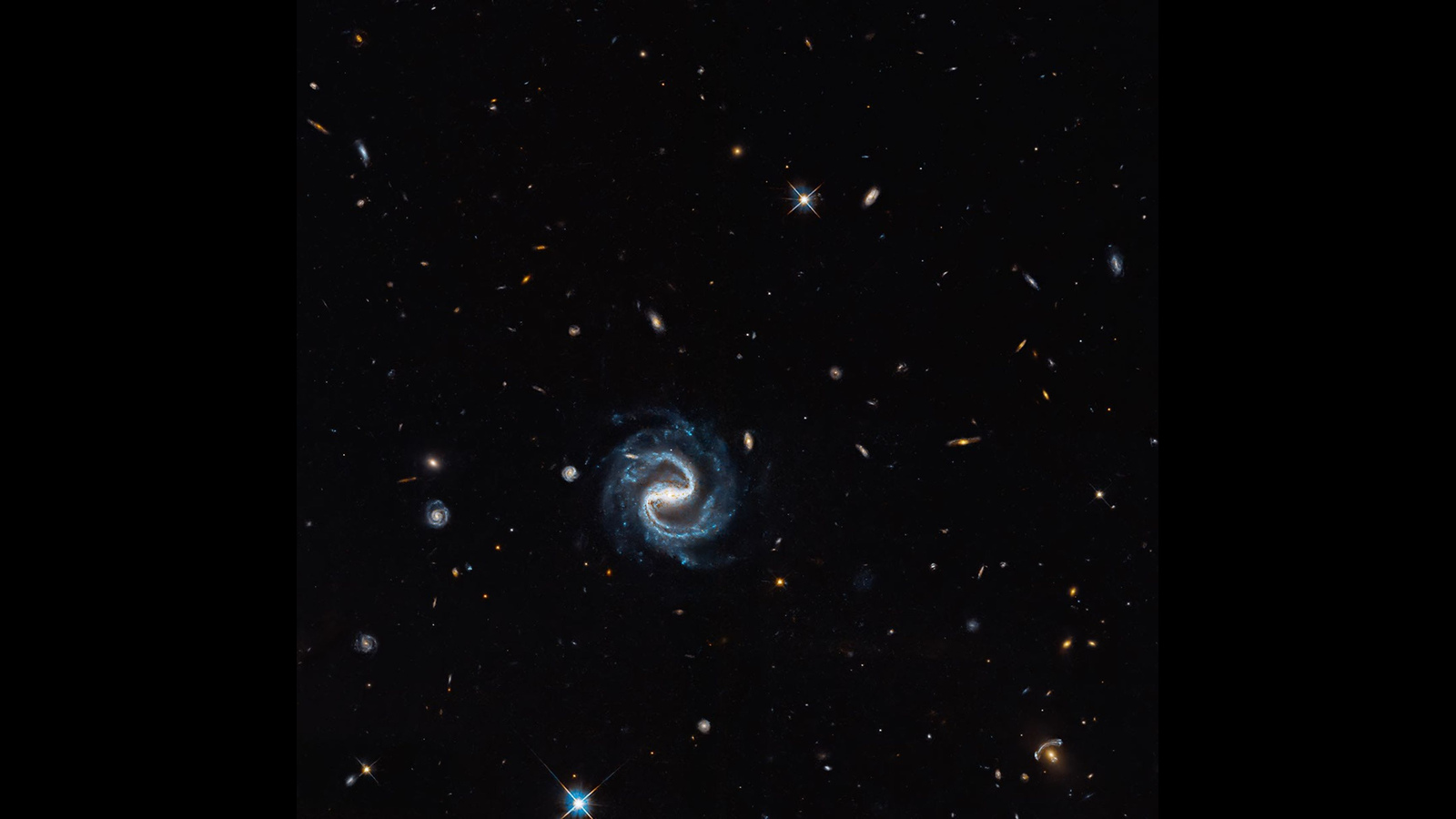 Imagen astronómica del Hubble: una galaxia espiral de brazos azules y, en el sector inferior derecho, los arcos de una galaxia distante deformada por una lente gravitacional. Difundida por NASA el 22 de octubre de 2026; fecha de captura no indicada. Observación completa presentada en un lienzo horizontal negro con bandas laterales; sin recorte de la imagen astronómica.