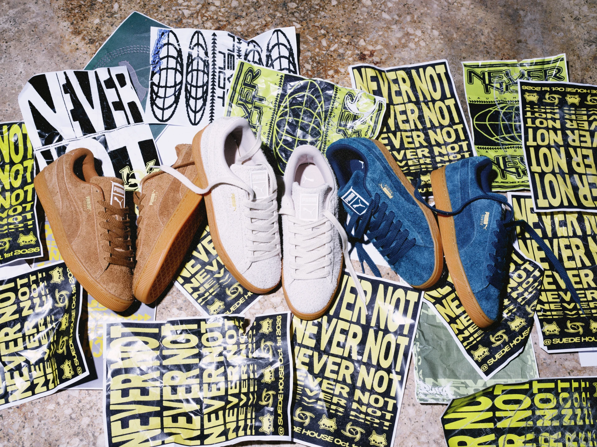 Tres pares de Puma Suede Forever en marrón, gris claro y azul petróleo sobre afiches Never Not Suede. Fotografía de producto del lanzamiento internacional del 1 de octubre de 2026.